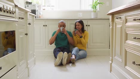 Happy-biracial-couple-sitting-on-kitchen's-floor,-drinking-coffee-and-talking