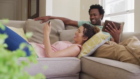 Glückliches-Gemischtrassiges-Paar,-Das-Sich-Mit-Smartphone-Und-Tablet-Auf-Dem-Sofa-Entspannt