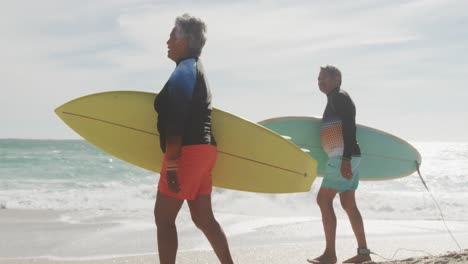 Side-view-of-happy-senior-hispanic-couple-walking-on-beach-with-surfboards