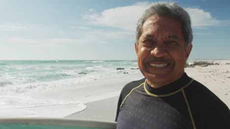 Portrait-of-happy-senior-hispanic-man-walking-on-beach-with-surfboard