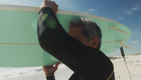 Happy-senior-hispanic-man-walking-on-beach-with-surfboard
