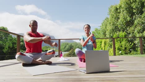 Happy-biracial-couple-on-terrace-exercising,-stretching-with-online-lessons
