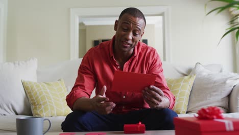 Happy-and-surprised-biracial-man-on-sofa,-having-video-call-and-reading-valentine's-card