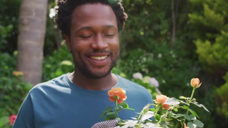 Portrait-of-happy-biracial-man-smelling-roses-in-garden