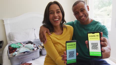 Happy-biracial-couple-sitting-on-bed-and-showing-smartphones-with-covid-19-passports-on-screens