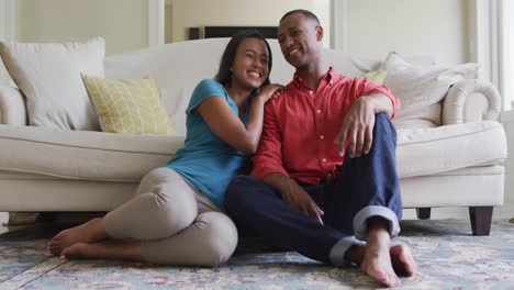 Happy-biracial-couple-sitting-on-floor-in-living-room,-embracing-and-looking-at-camera