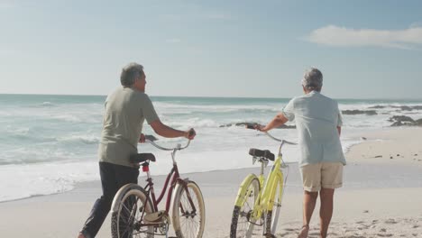 Back-view-of-hispanic-senior-couple-walking-with-bikes-on-beach-at-sunset