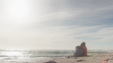 Hispanic-senior-couple-sitting-and-embracing-on-beach-at-sunset