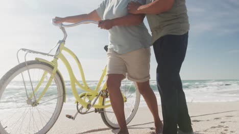Midsection-of-hispanic-senior-couple-walking-with-bikes-on-beach-at-sunset