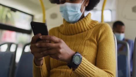 African-american-businesswoman-with-face-mask-using-smartphone-and-sitting-in-bus