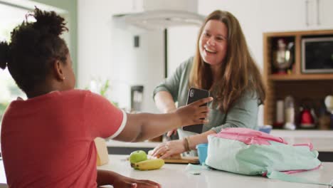 Happy-caucasian-woman-and-her-african-american-daughter-using-smartphone-in-kitchen