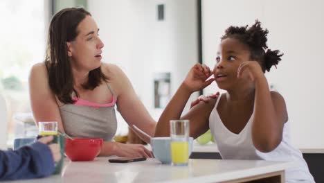 Happy-caucasian-woman-and-her-african-american-daughter-eating-breakfast-in-kitchen