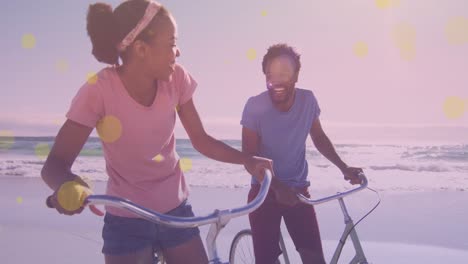 Gelbe-Lichtpunkte-Vor-Einem-Afroamerikanischen-Paar,-Das-Auf-Seinen-Fahrrädern-Sitzt-Und-Am-Strand-Lächelt