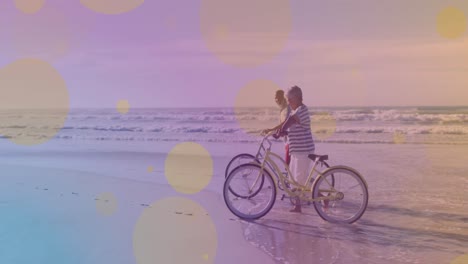 Gelbe-Lichtpunkte-Vor-Afroamerikanischem-Seniorenpaar-Mit-Fahrrädern,-Die-Gemeinsam-Am-Strand-Spazieren-Gehen