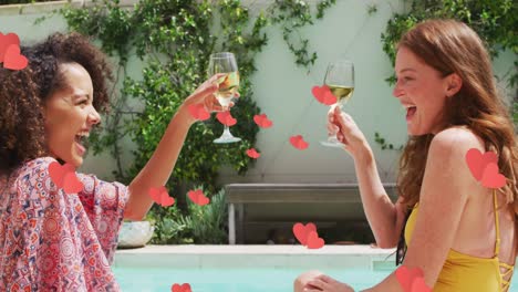 Multiple-red-hearts-floating-against-two-diverse-female-friends-toasting-drinks-by-the-pool