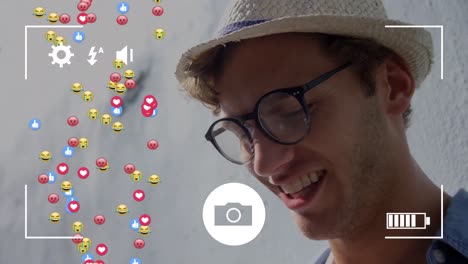 Camera-interface-over-multiple-face-emojis-and-icons-floating-against-caucasian-man-smiling