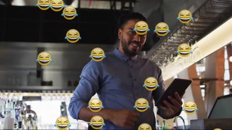Múltiples-Emojis-De-Caras-Risueñas-Contra-Un-Barman-Afroamericano-Usando-Una-Tableta-Digital-En-Un-Bar