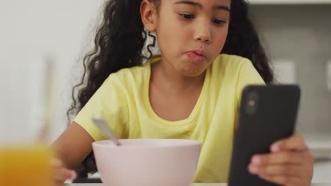 Fröhliches-Gemischtrassiges-Mädchen-Am-Tisch,-Das-Frühstück-Isst-Und-Sein-Smartphone-Benutzt
