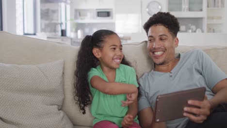 Happy-biracial-father-and-daughter-sitting-on-sofa-using-tablet