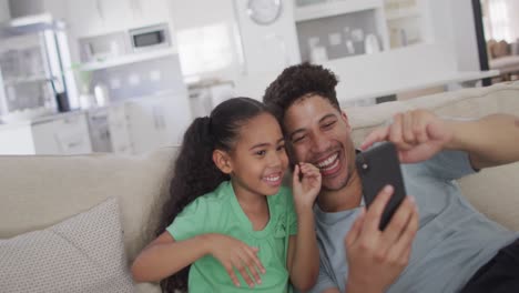 Happy-biracial-father-and-daughter-sitting-on-sofa-using-smartphone