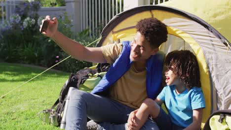 Happy-biracial-man-and-his-son-taking-selfies-using-smartphone-in-garden