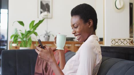 Happy-african-american-woman-sitting-on-sofa-in-living-room,-using-smartphone