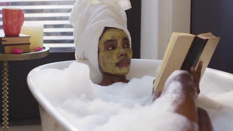 Afroamerikanische-Frau-Mit-Handtuch-Und-Maske,-Die-Ein-Bad-Nimmt-Und-Im-Badezimmer-Ein-Buch-Liest