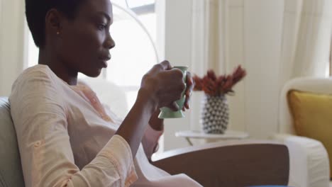Happy-african-american-woman-sitting-on-armchair-in-living-room,-drinking-coffee