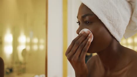 Smiling-african-american-woman-with-towel-using-pad-on-her-face-and-watching-in-mirror-in-bathroom