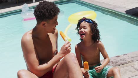 Happy-biracial-man-and-his-son-eating-ice-cream-by-swimming-pool-in-garden