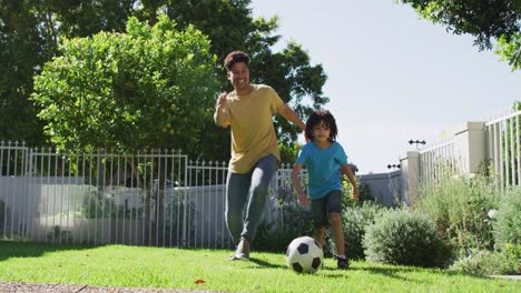 Happy-biracial-man-and-his-son-playing-football-in-garden