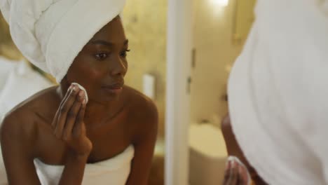 Smiling-african-american-woman-with-towel-watching-in-mirror-and-using-pad-on-her-face-in-bathroom