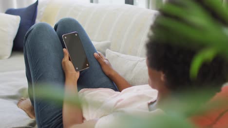 Happy-african-american-woman-sitting-on-sofa,-using-smartphone-with-copy-space