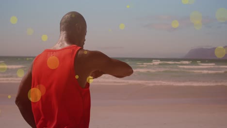 Yellow-spots-floating-against-african-american-fit-man-using-smartwatch-at-the-beach
