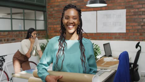 Portrait-of-happy-african-american-female-architect-holding-architectural-blueprints-at-office