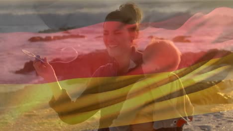 Animation-Der-Flagge-Deutschlands-über-Einer-Glücklichen-Kaukasischen-Mutter-Mit-Kind,-Das-Am-Strand-Seifenblasen-Bläst