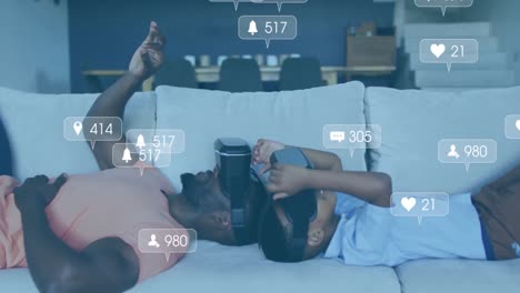 Animation-of-social-media-icons-and-numbers-over-african-american-father-and-son-using-vr-headset