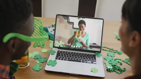 Smiling-african-american-woman-with-beer-wearing-clover-shape-band-on-video-call-on-laptop