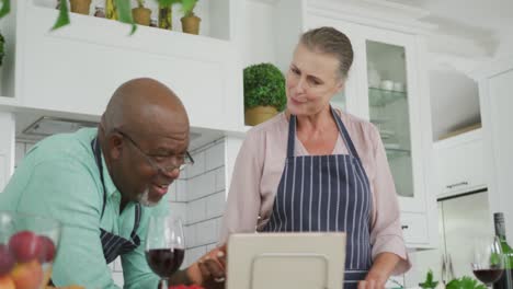 Lächelndes-älteres,-Vielfältiges-Paar,-Das-Blaue-Schürzen-Trägt-Und-In-Der-Küche-Ein-Tablet-Benutzt