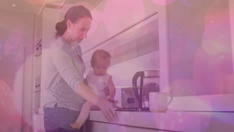 Animation-of-pink-spots-of-light-over-caucasian-woman-with-baby-in-kitchen