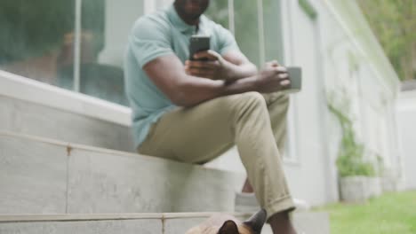 Glücklicher-Afroamerikanischer-Mann,-Der-Mit-Smartphone-Sitzt-Und-Kaffee-Im-Garten-Trinkt