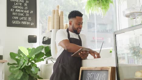 African-american-male-cafe-owner-using-laptop-and-tablet-at-cafe