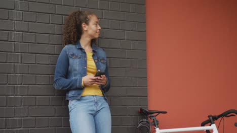 Happy-biracial-woman-in-city,-next-to-bike-using-smartphone