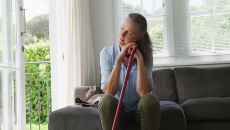 Senior-caucasian-woman-wearing-blue-shirt-and-sitting-with-cane-and-cat-in-living-room
