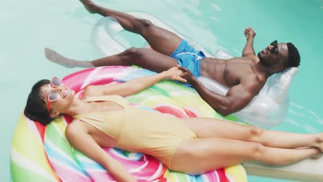 Happy-diverse-couple-wearing-swimming-suits-with-inflatables-at-swimming-pool-in-garden
