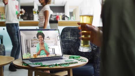 Mujer-Afroamericana-Sonriente-Con-Cerveza-Usando-Una-Banda-En-Forma-De-Trébol-En-Videollamada-En-Una-Computadora-Portátil