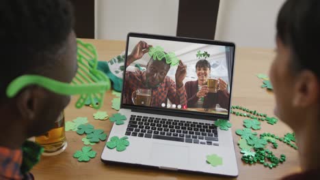 Smiling-diverse-couple-with-beer-wearing-clover-shape-items-on-video-call-on-laptop