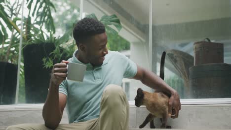 Happy-african-american-man-sitting-with-cat-and-drinking-coffee-in-garden