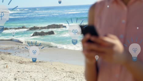 Animation-of-bulb-icons-over-midsection-of-biracial-woman-using-smartphone-at-beach