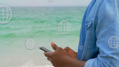 Animation-Von-Globen-über-Dem-Mittelteil-Eines-Afroamerikanischen-Mannes,-Der-Am-Strand-Sein-Smartphone-Benutzt